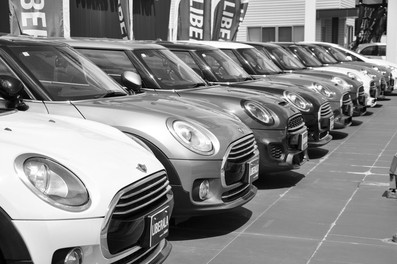 our-story-02 Black and white photo of a row of Mini Cooper cars parked outdoors in a car dealership.